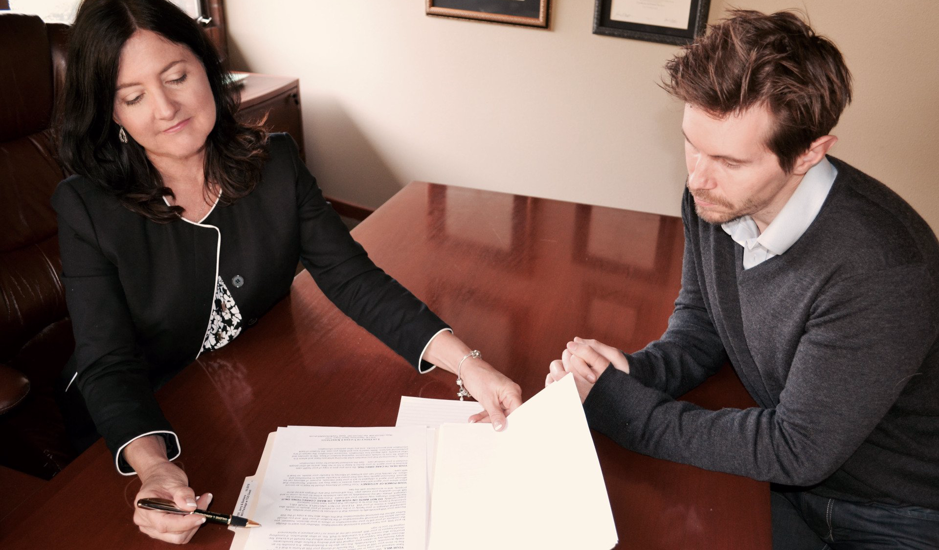 Woman in black blazer points to documents as man in sweater looks on at a desk.