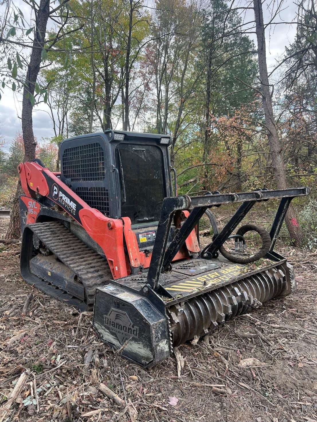 Orange and black skid steer with forestry mulcher in a wooded area.