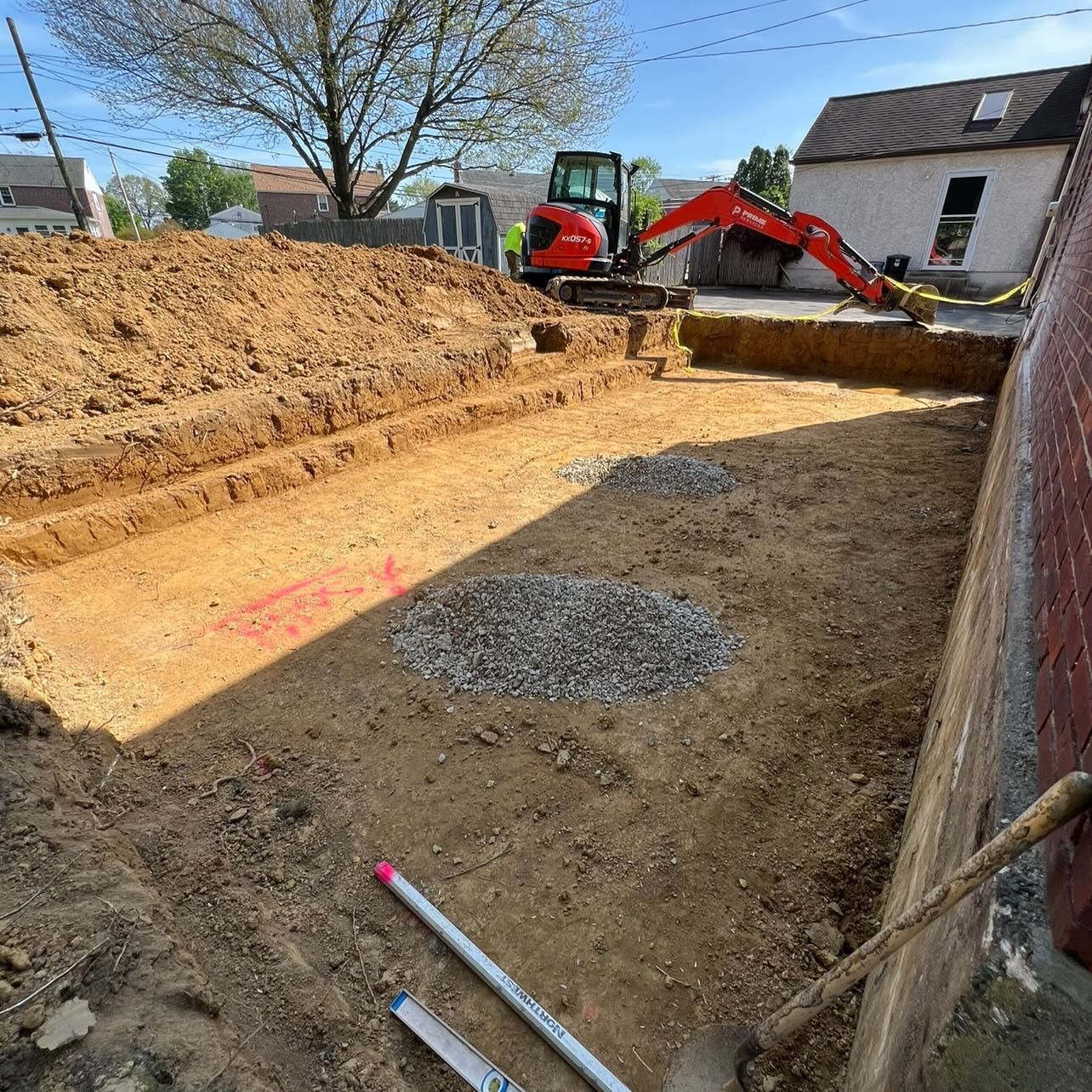 Construction site with an excavator digging trenches in a dirt lot next to a brick building.