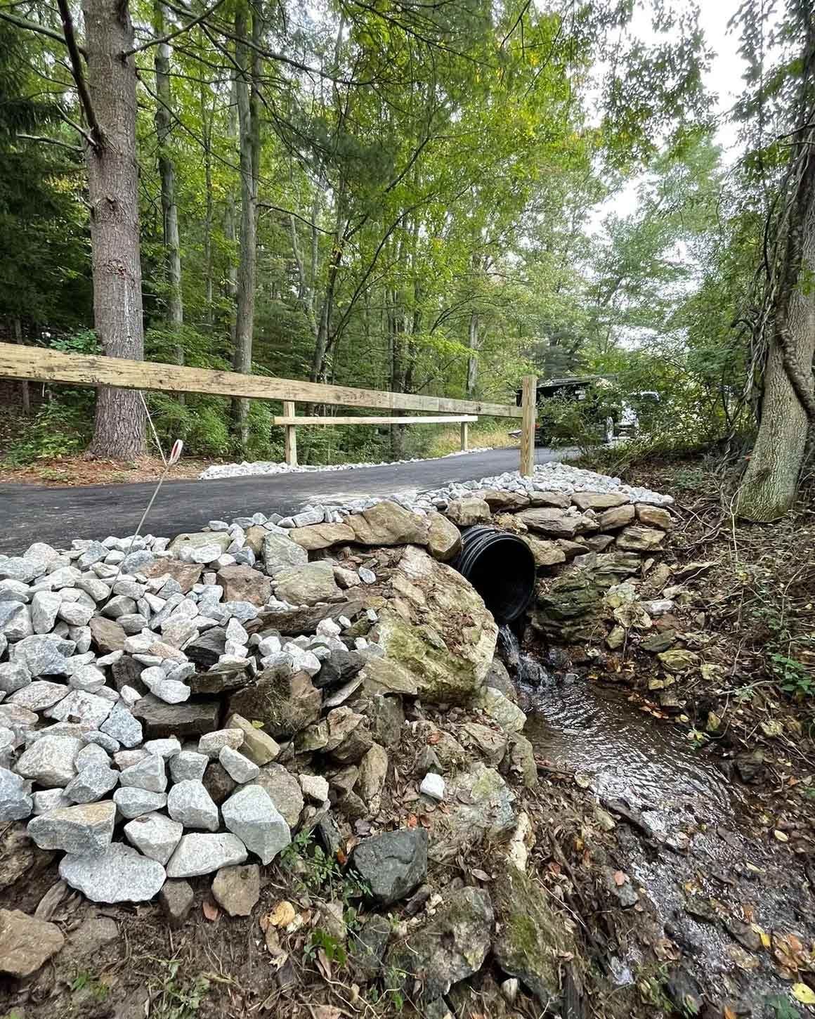 Stone bridge with culvert carrying water under a paved path with wooden rail fence; forest backdrop.