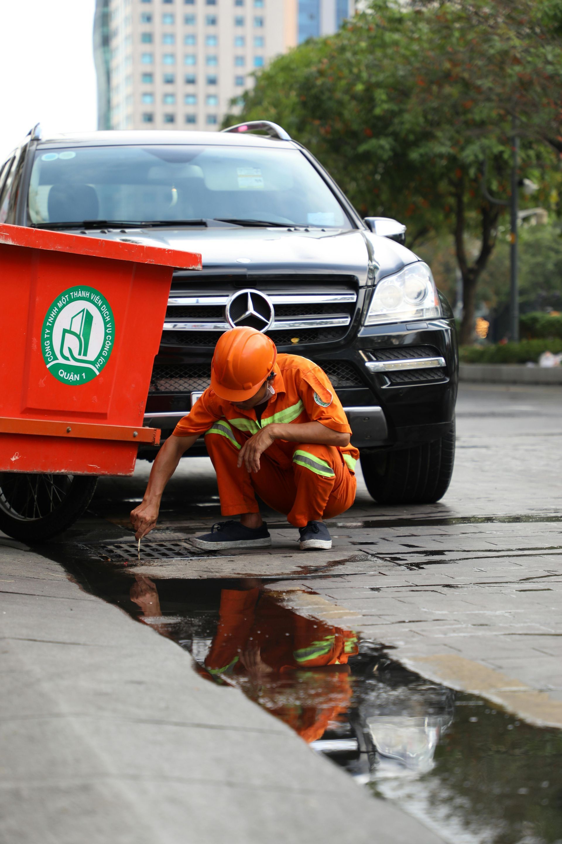 Worker in orange uniform crouches by a wet curb beside a red cart and black SUV.