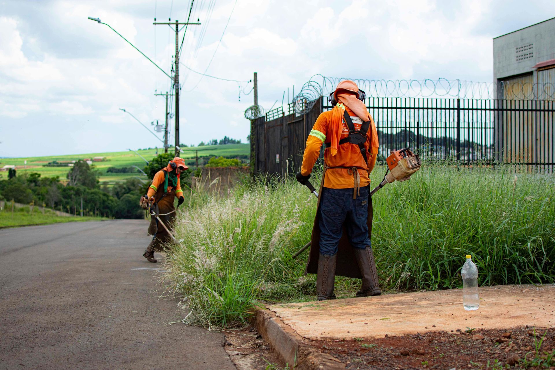 Workers in orange safety gear trimming weeds along a roadside near a fence and field.