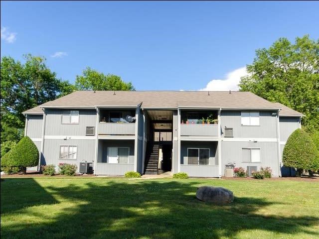 Gray two-story apartment building with a central staircase, surrounded by green grass and trees on a sunny day.