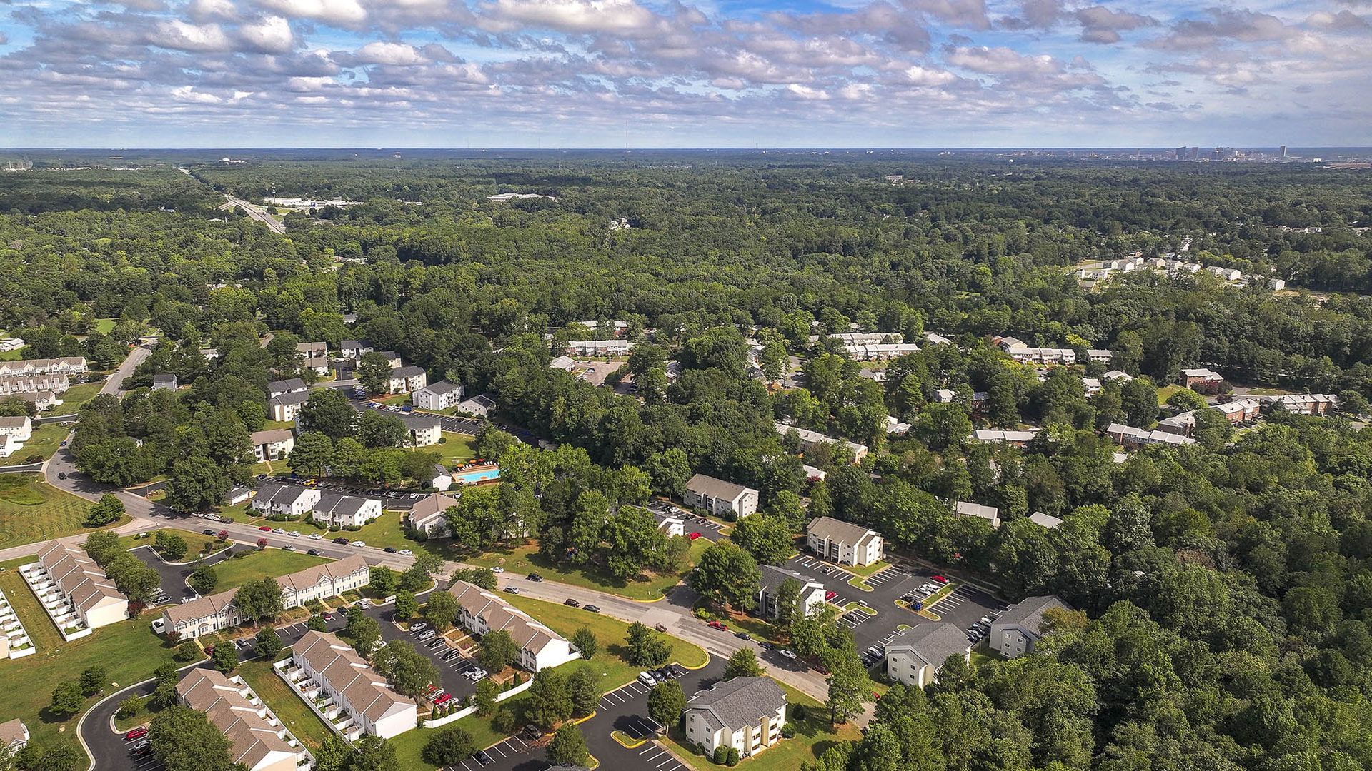 Aerial view of a suburban neighborhood with houses, roads, and a vast green forest under a partly cloudy sky.