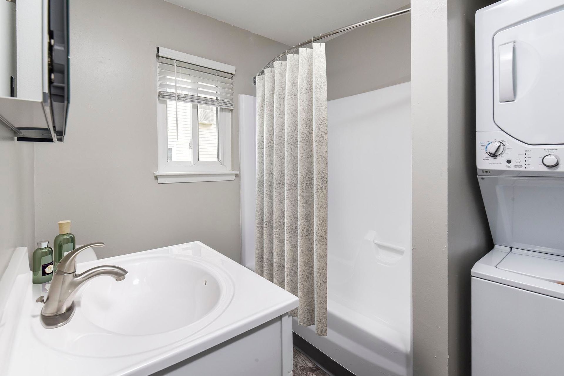 Bathroom with white sink, shower, and stacked washer/dryer; gray walls and window with blinds.