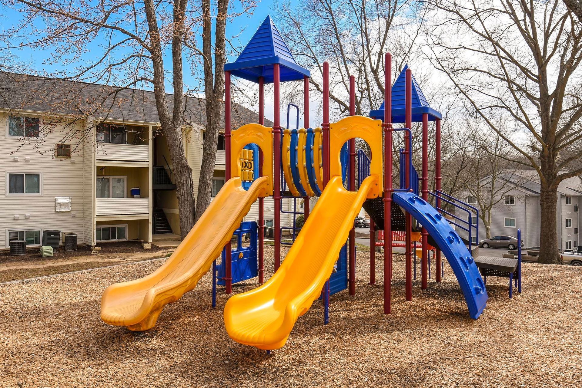 Playground with yellow and blue slides, red poles, and wood chip ground near apartment buildings.