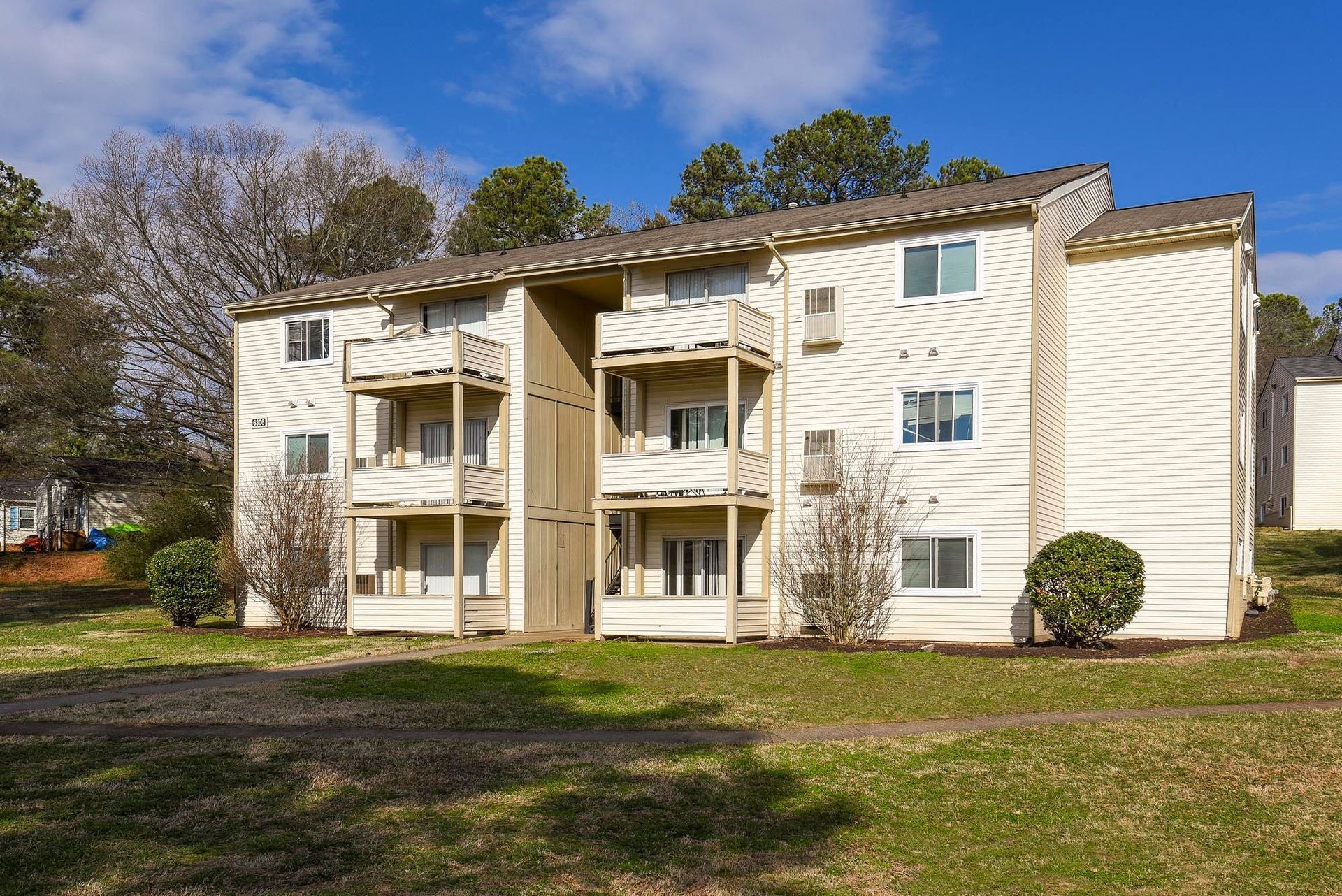 Two-story apartment building with balconies, beige siding, and brown roof. Situated on grassy lawn with trees in the background.