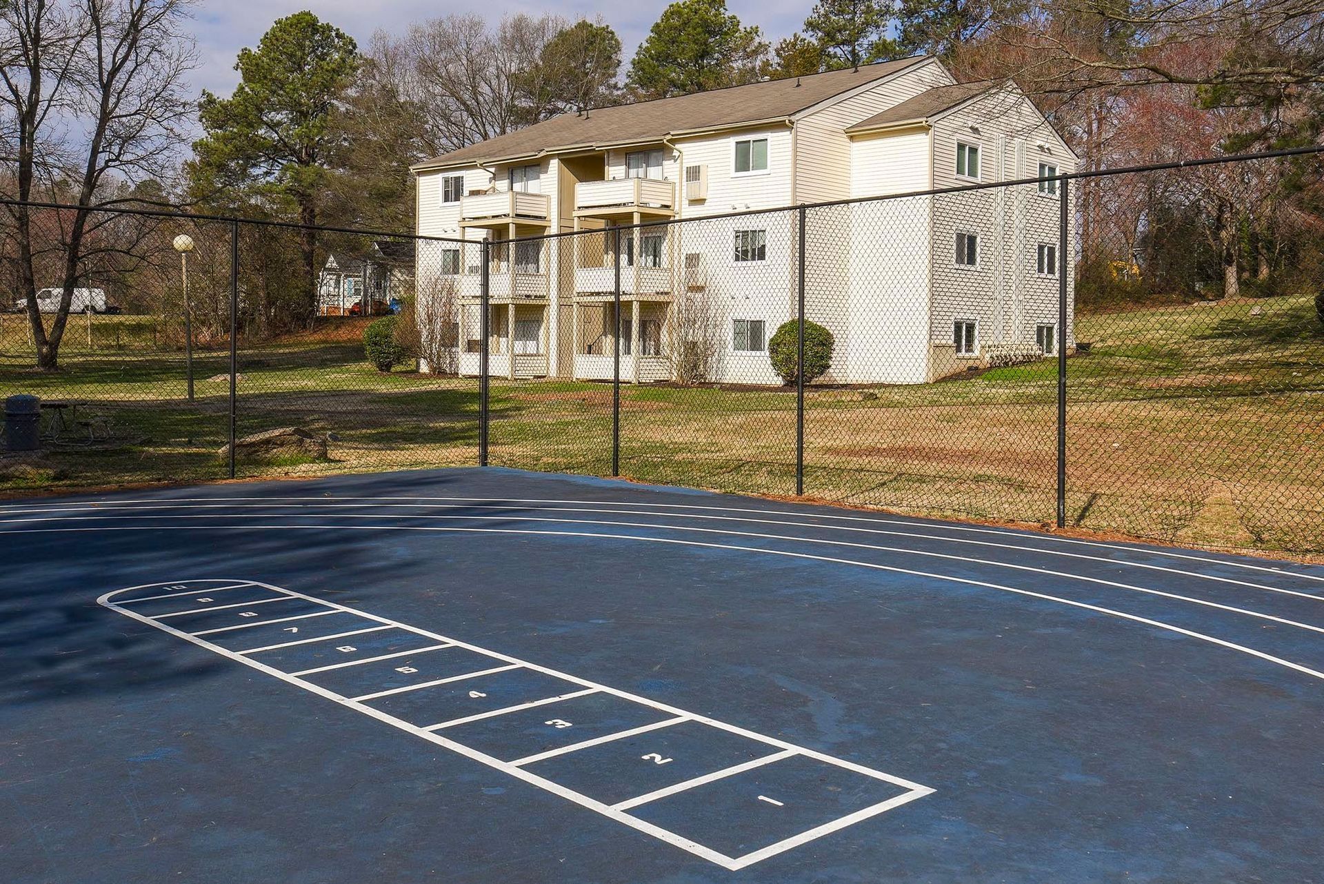 A blue asphalt playground with a hopscotch grid in front of a multi-story building behind a fence.