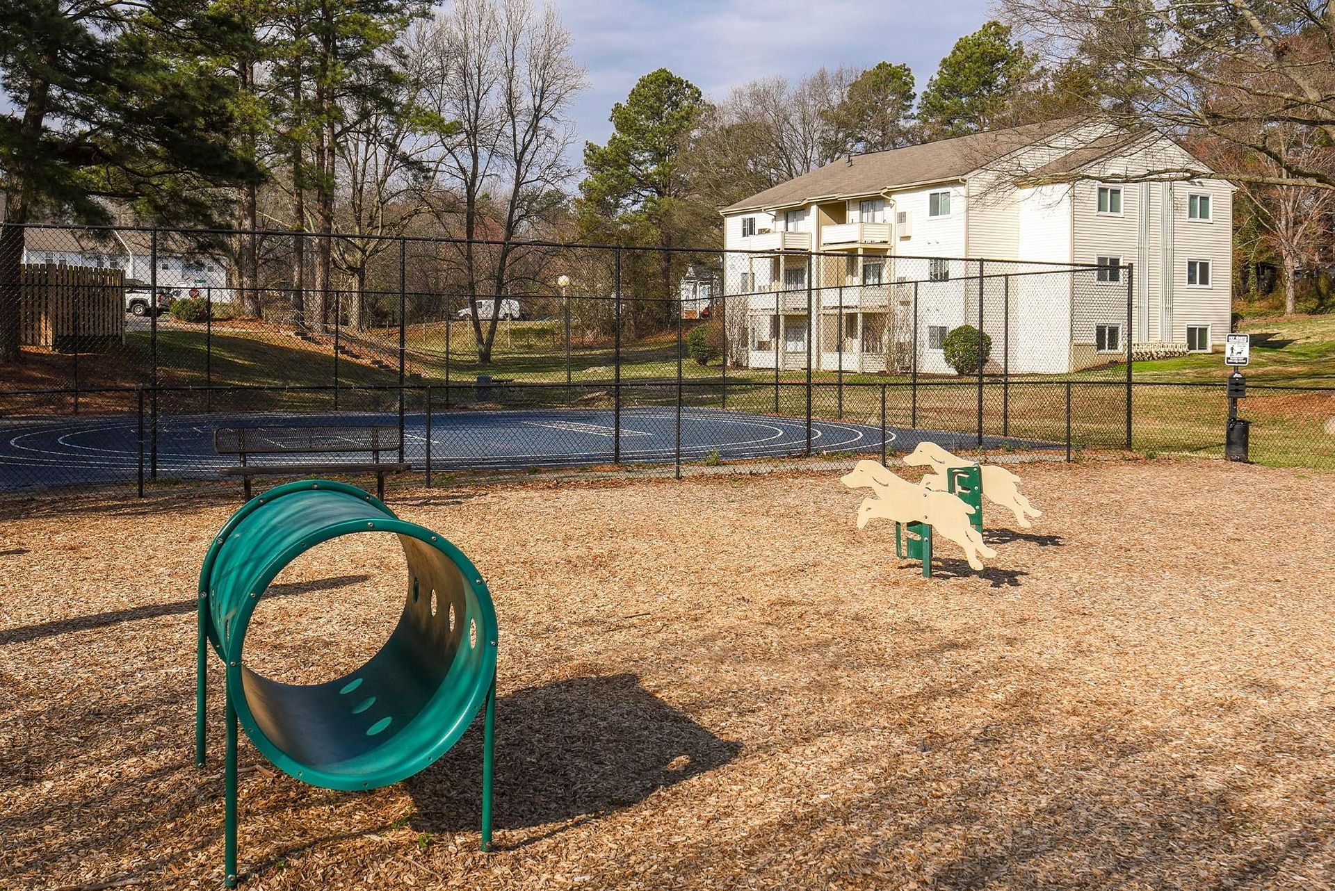 Dog park with tunnel and agility equipment; wood chip ground, fenced, apartment building background.