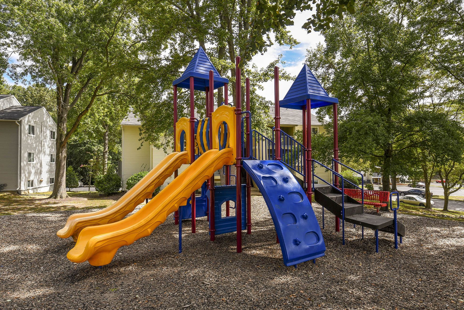 Playground with yellow slides, blue climbing wall, and dark mulch, surrounded by trees.