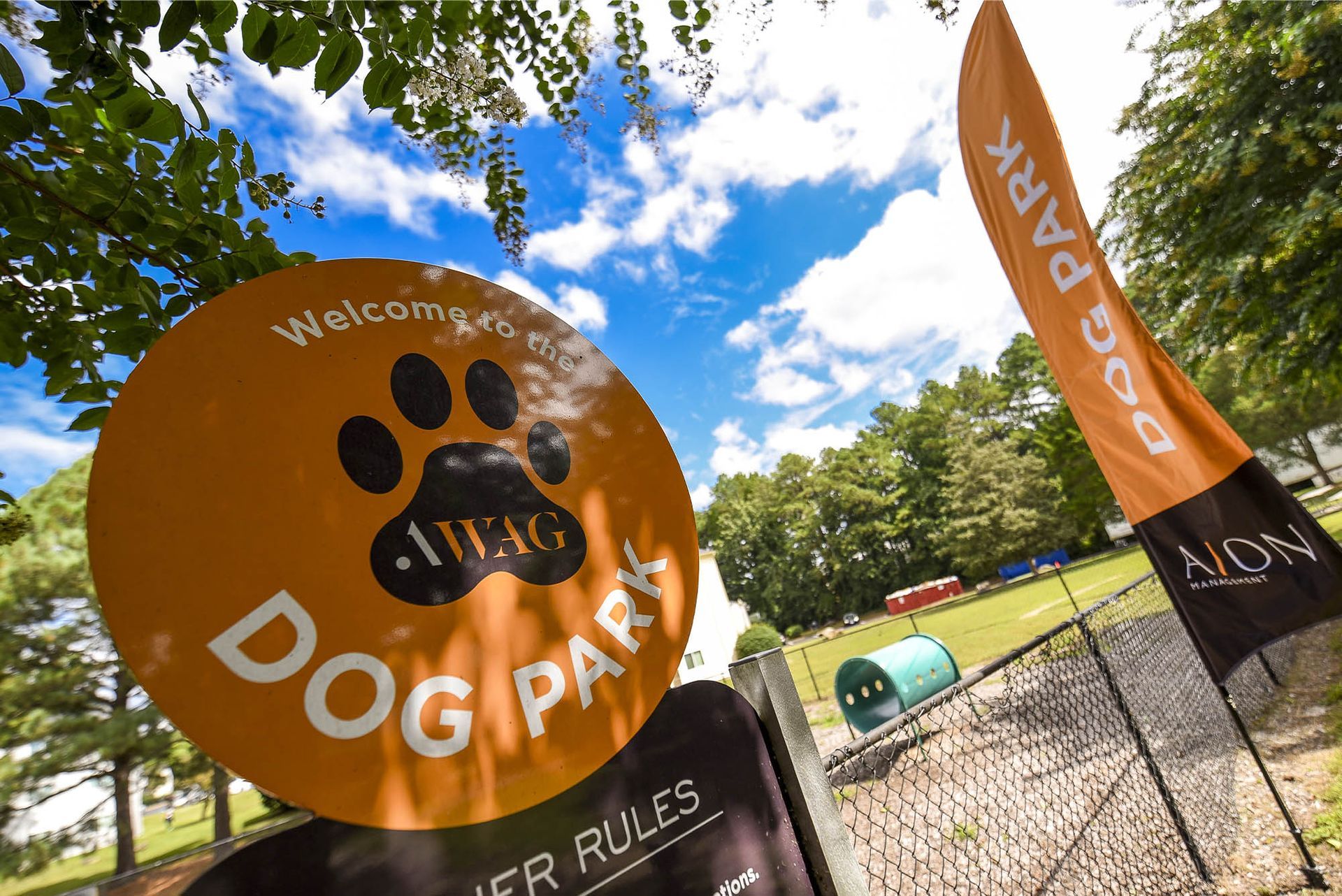 Orange sign welcoming to a dog park with a paw print. Orange flag on right reads 