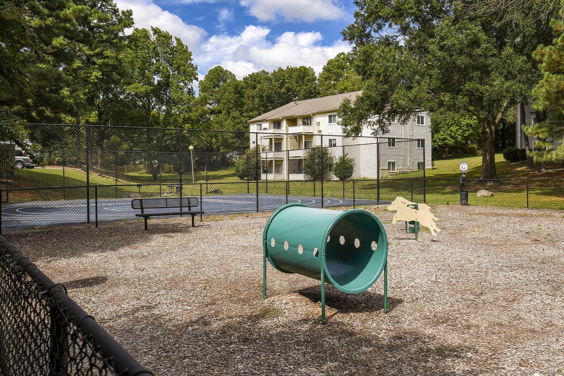 Dog park with tunnel, bench, and low fence, trees and apartment building in the background on a sunny day.