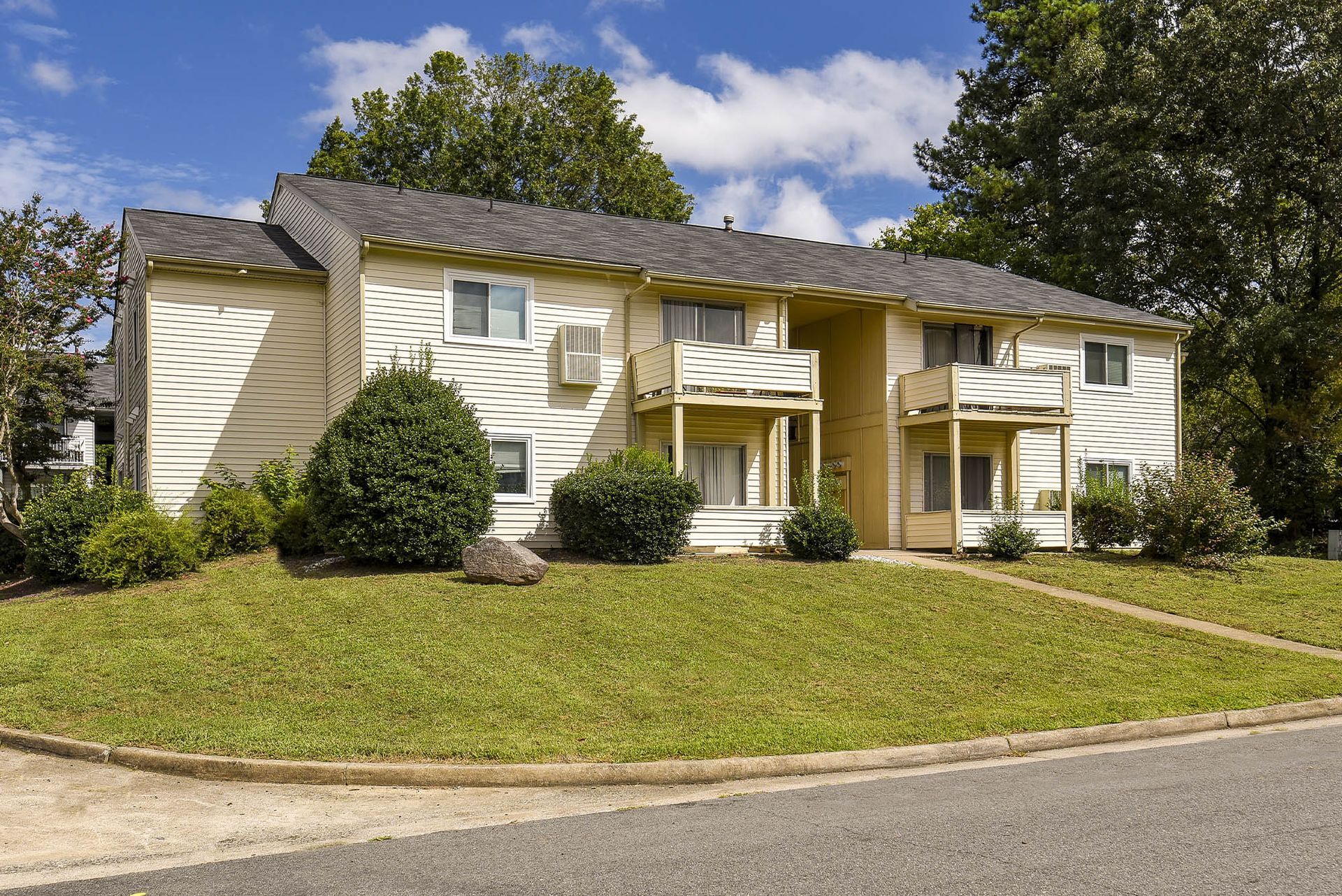 Two-story tan apartment building with balconies, set on a green lawn with trees.
