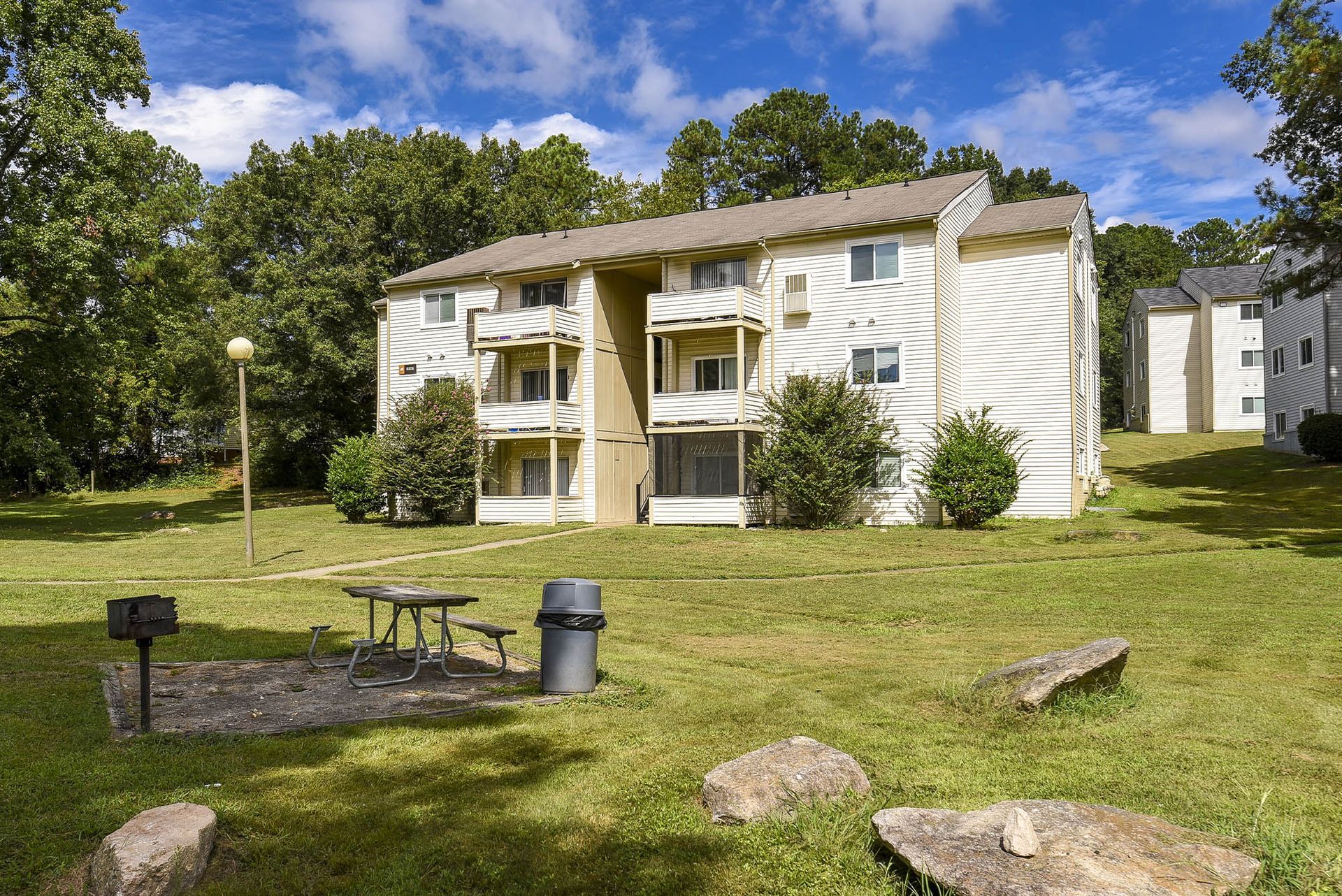 Apartment building with grass, trees, picnic table, grill, and garbage can on a sunny day.
