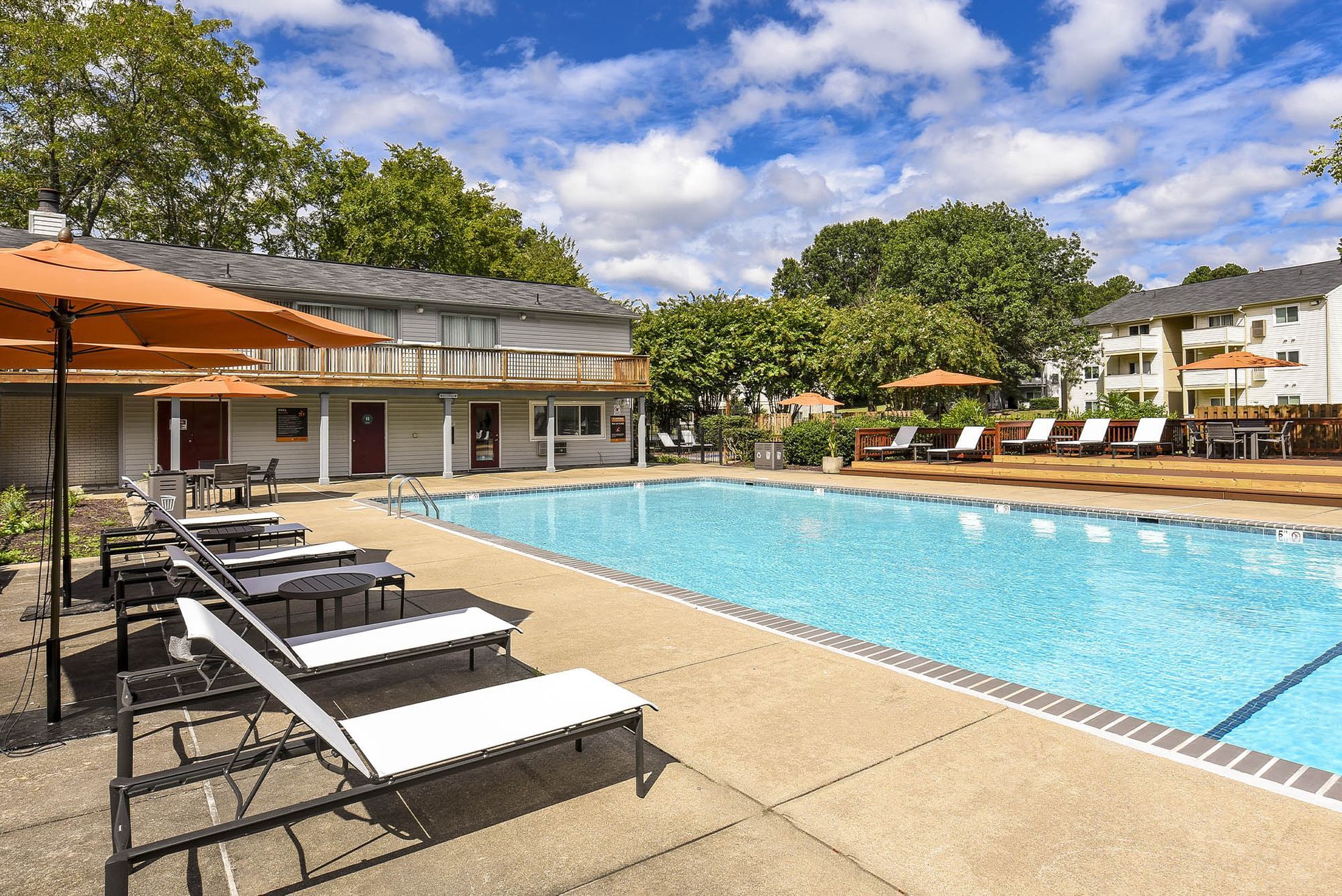 Swimming pool with lounge chairs, umbrellas, and apartments in the background under a blue sky.