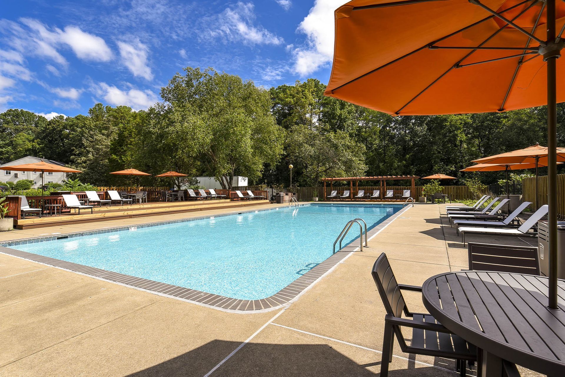 Swimming pool with lounge chairs, orange umbrellas, and a table with chairs. Blue sky and trees in the background.