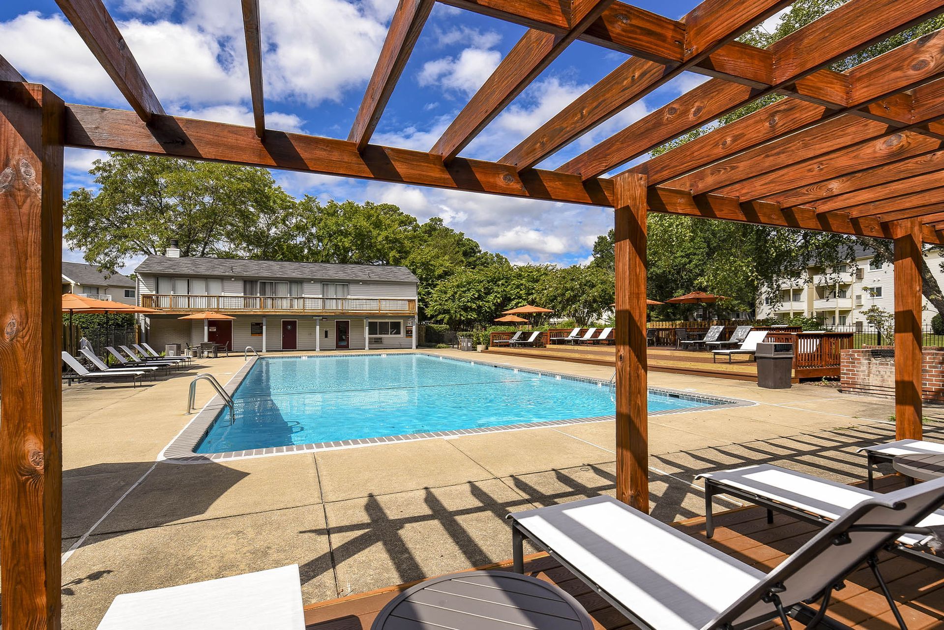 Pool with wooden pergola, lounge chairs, and buildings in the background under a blue sky.
