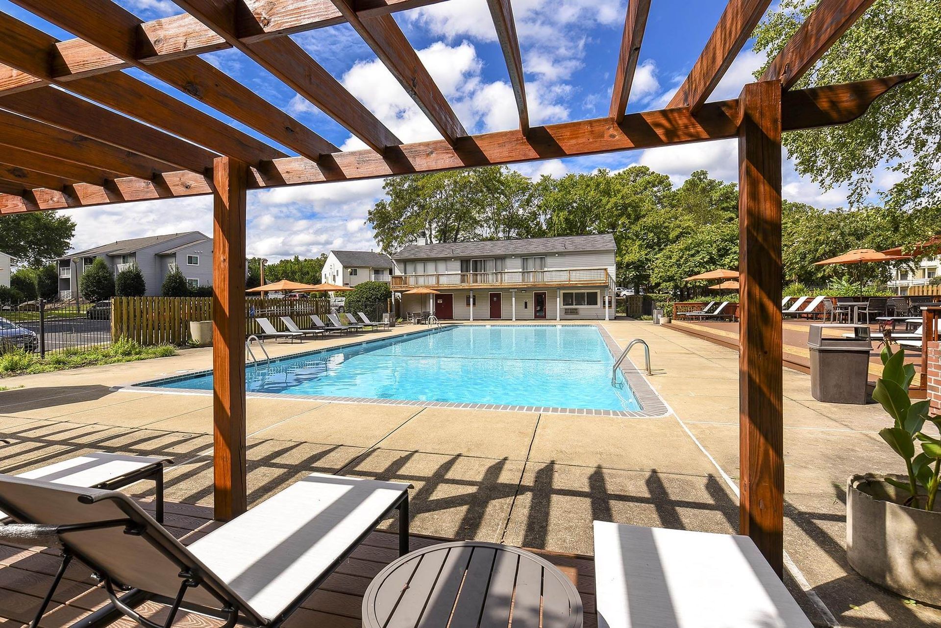 Pool area with wooden pergola, lounge chairs, and building in the background on a sunny day.