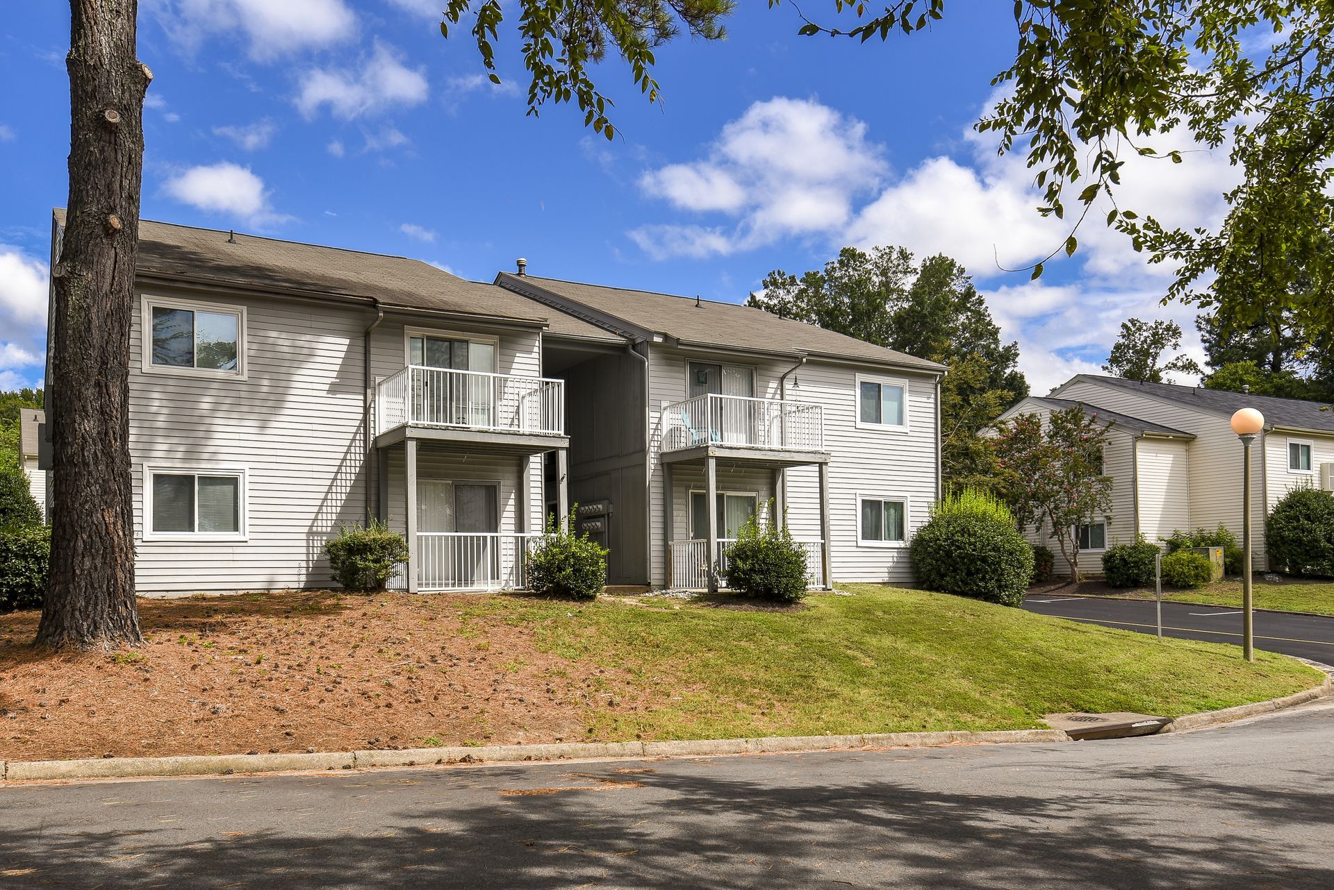 Two-story tan apartment building with balconies, set on a green lawn with trees.