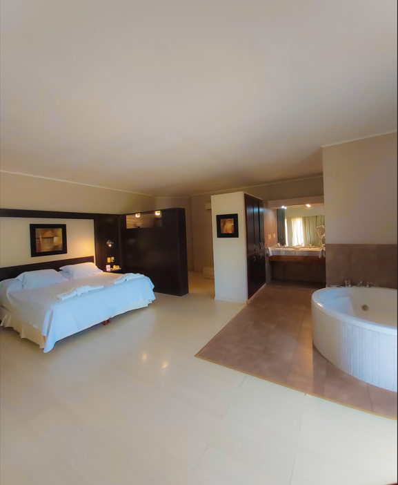 Una suite de hotel moderna y espaciosa que cuenta con una cama blanca a la izquierda y una bañera de hidromasaje redonda blanca sobre un suelo de baldosas a la derecha.