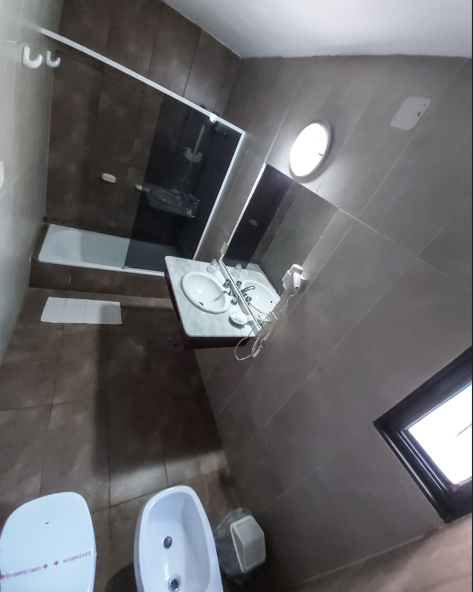 Un baño moderno con paredes revestidas de azulejos oscuros, una ducha con mampara de cristal, un tocador suspendido con lavabo y un bidé.