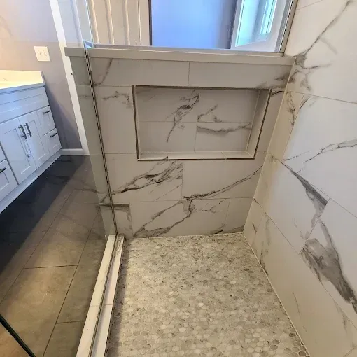 Shower with marble-look tile, a built-in niche, and a mosaic-tiled floor. White cabinets and window visible.