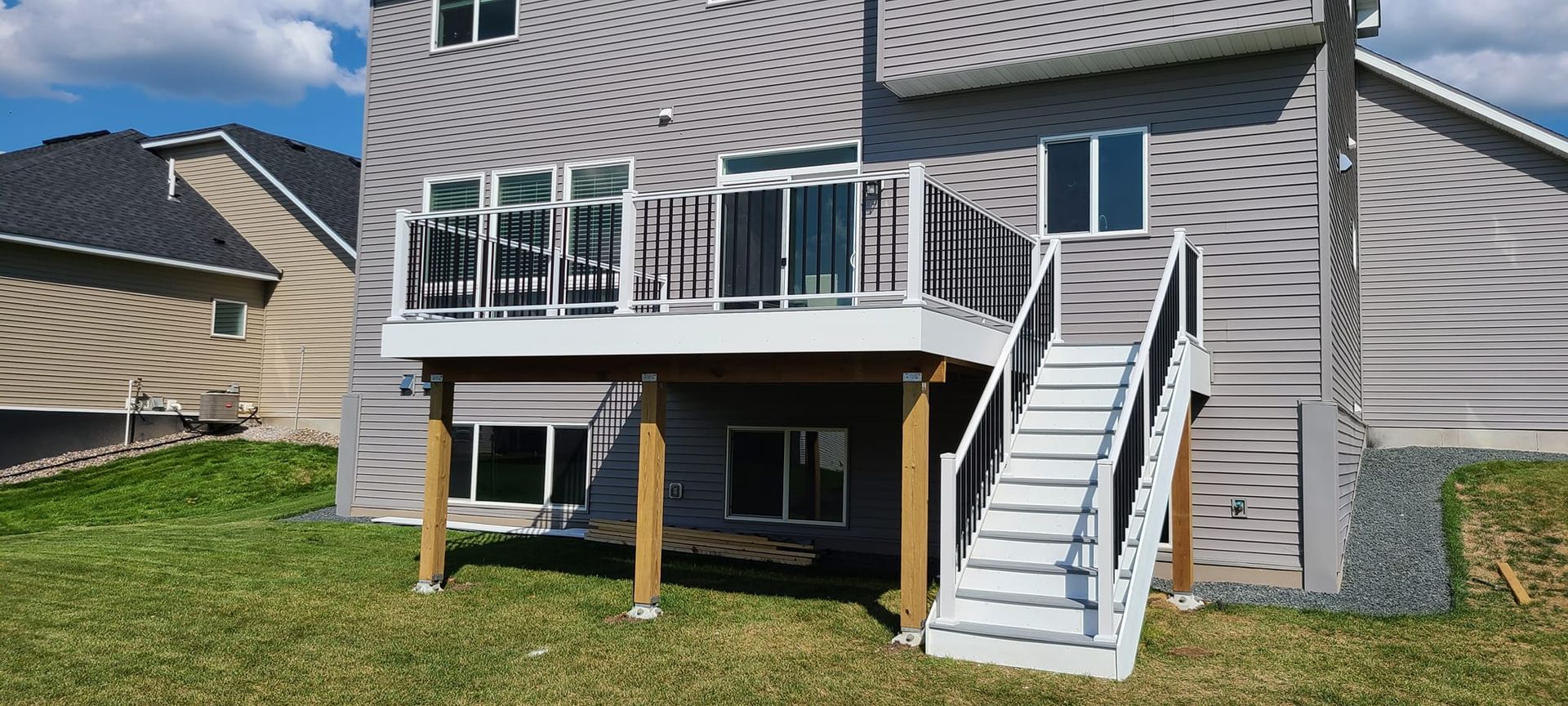 A gray house with a white deck and stairs, surrounded by green grass and other houses in the background.