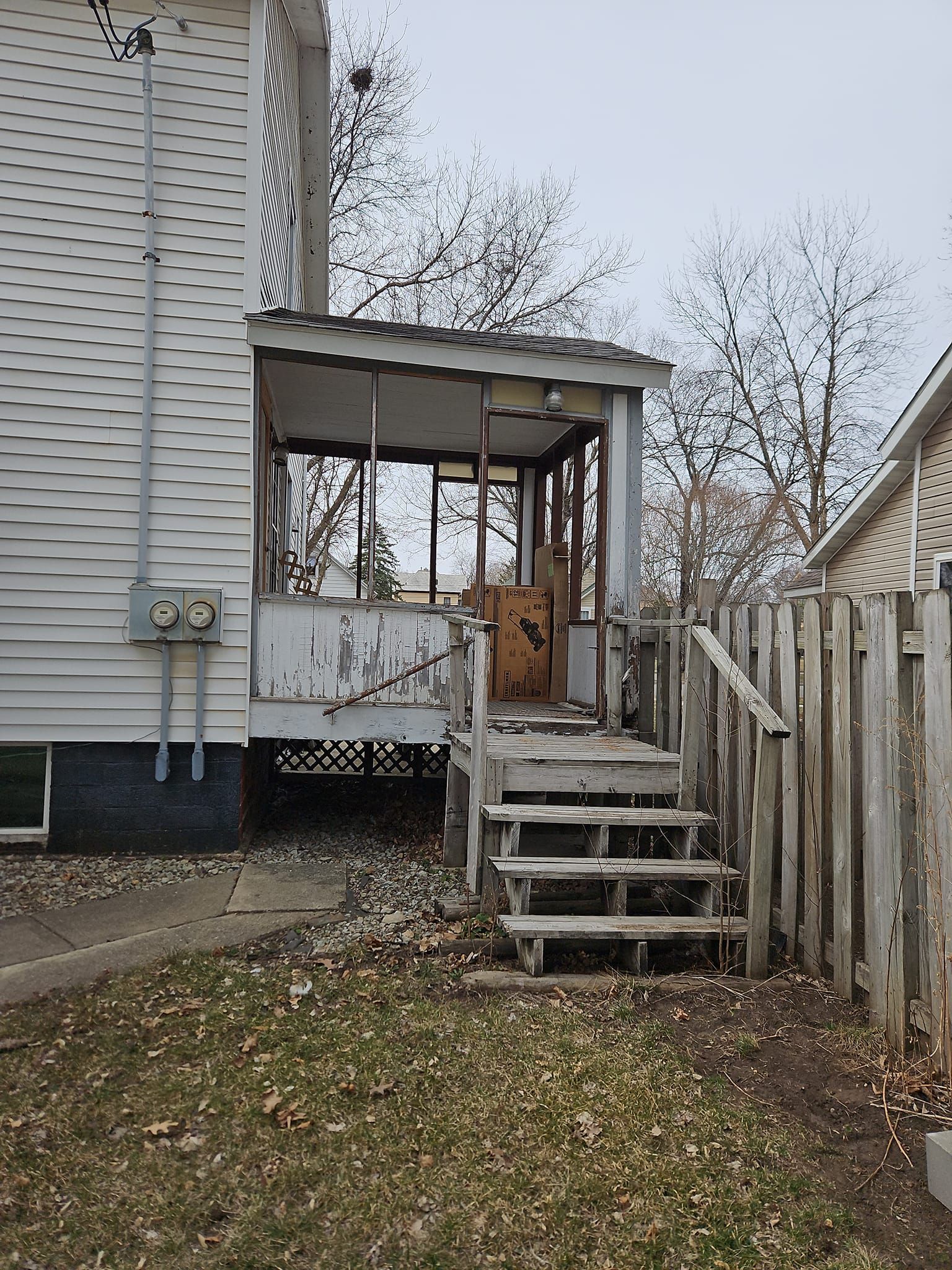 A weathered porch with stairs. House siding to the left, a wooden fence to the right, and a yard covered in leaves.