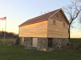 Barn-like structure with light wood siding and stone foundation, American flag flying, and sunset background.