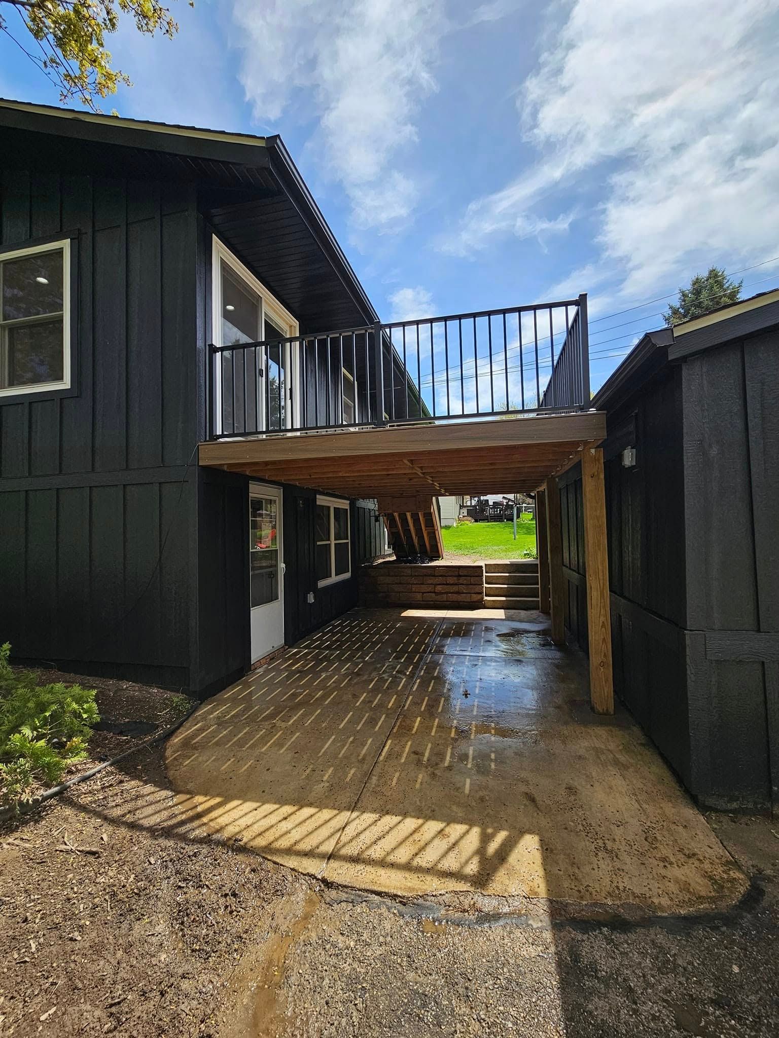 Black house with a wooden deck, a covered concrete patio, and stairs leading to a grassy yard.