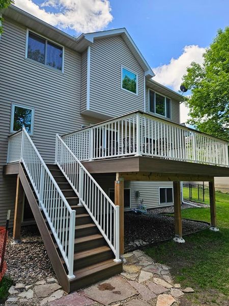 Two-story house with a deck and stairs. Tan siding, white railings, brown deck. Green lawn, blue sky.
