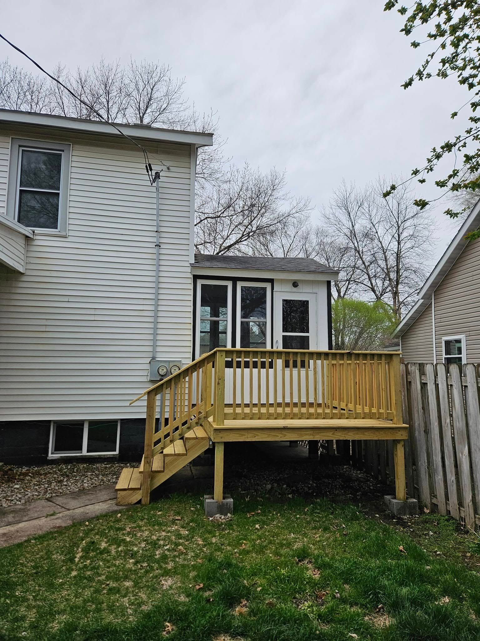 Backyard deck with stairs attached to a small white house and surrounded by grass and a wooden fence.
