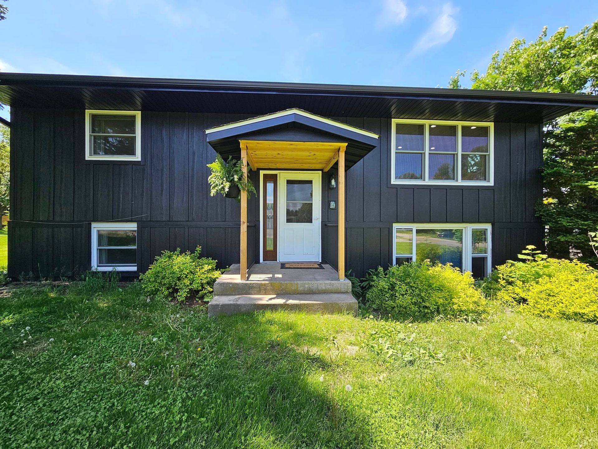 Black-sided split-level house with white trim, a small covered porch, and a green lawn under a blue sky.