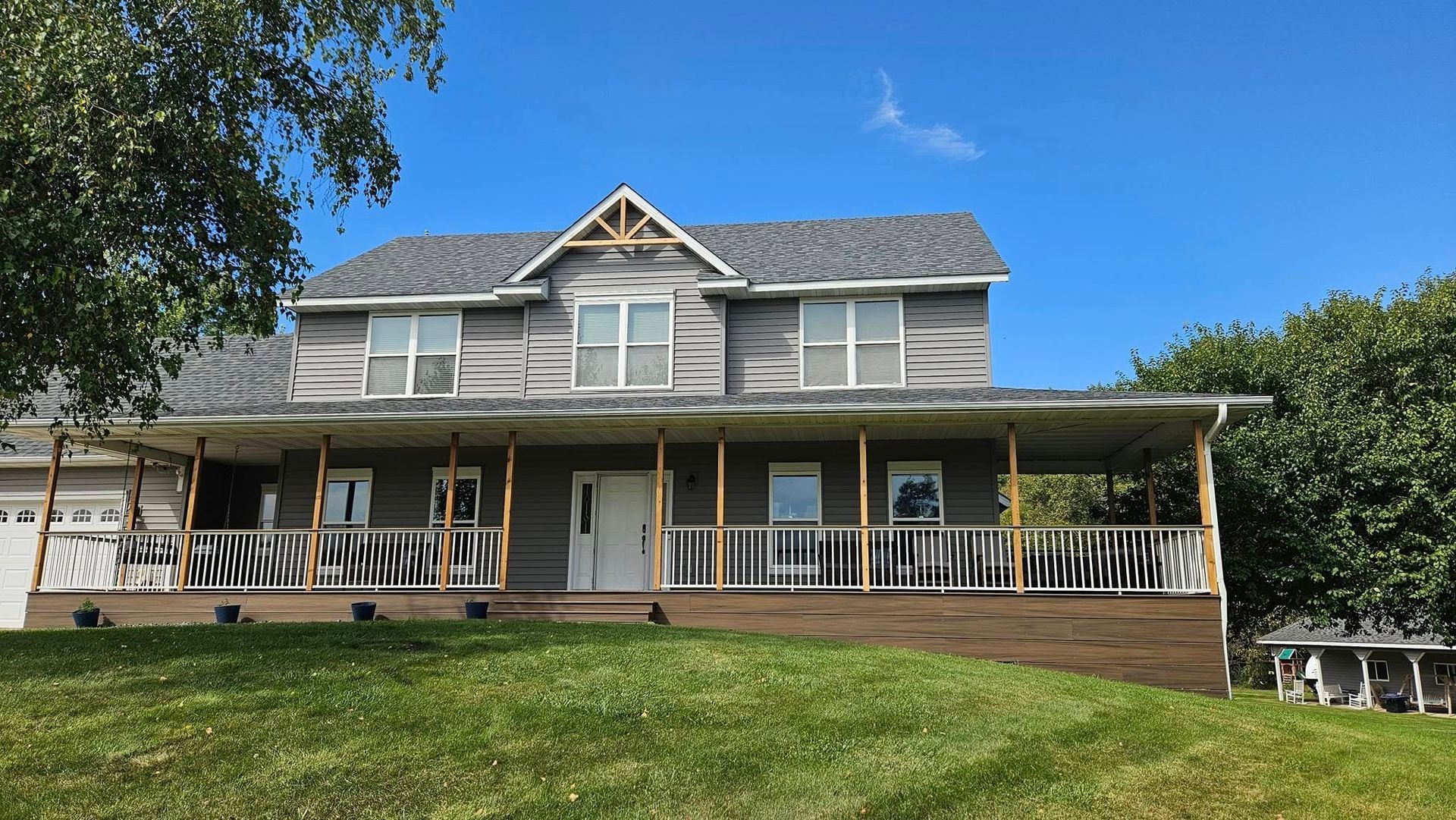 Two-story grey house with a wrap-around porch on a grassy hill under a blue sky.