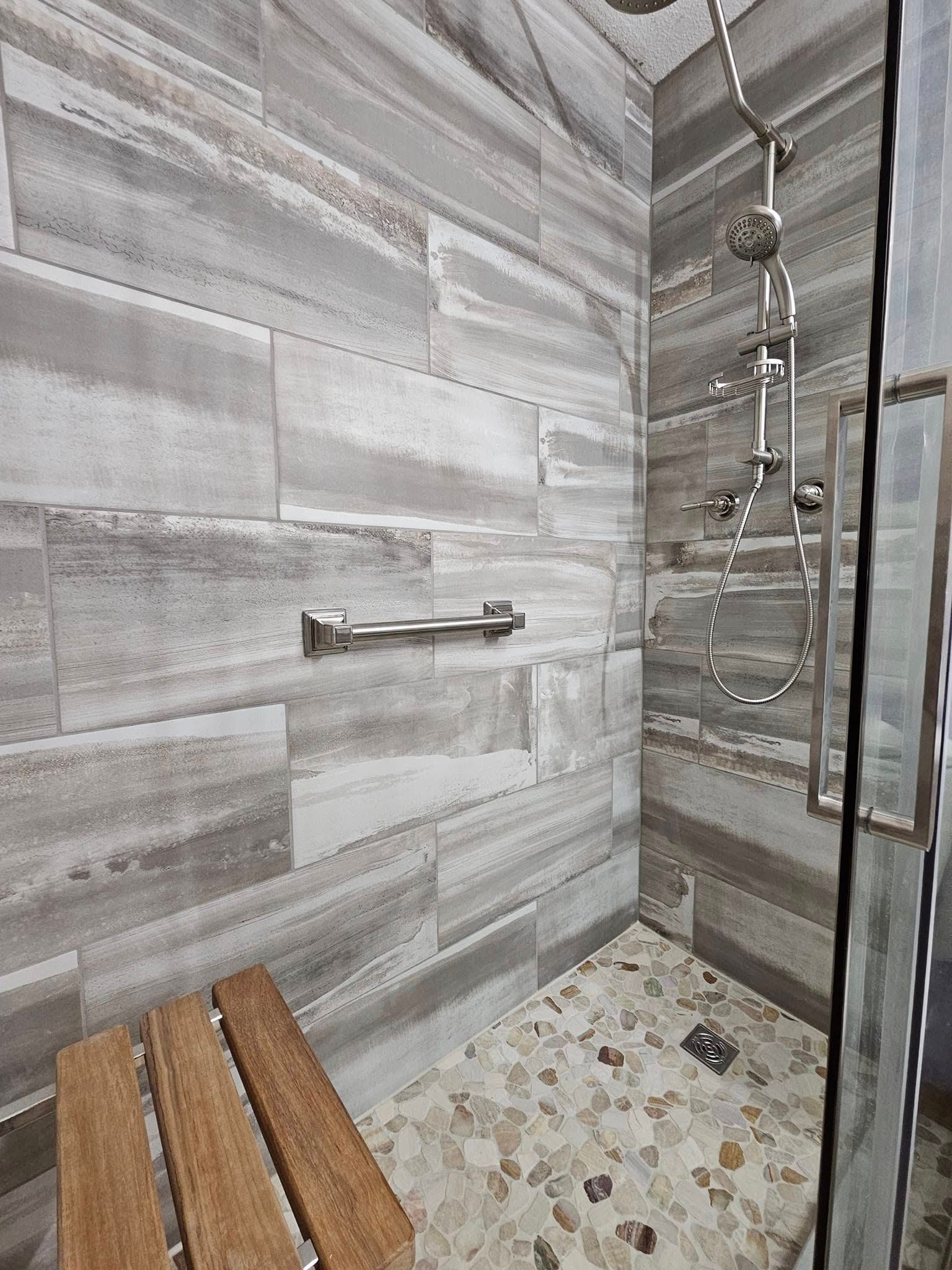 Accessible shower with stone floor, gray and white tile walls, grab bar, wooden bench, and glass door.