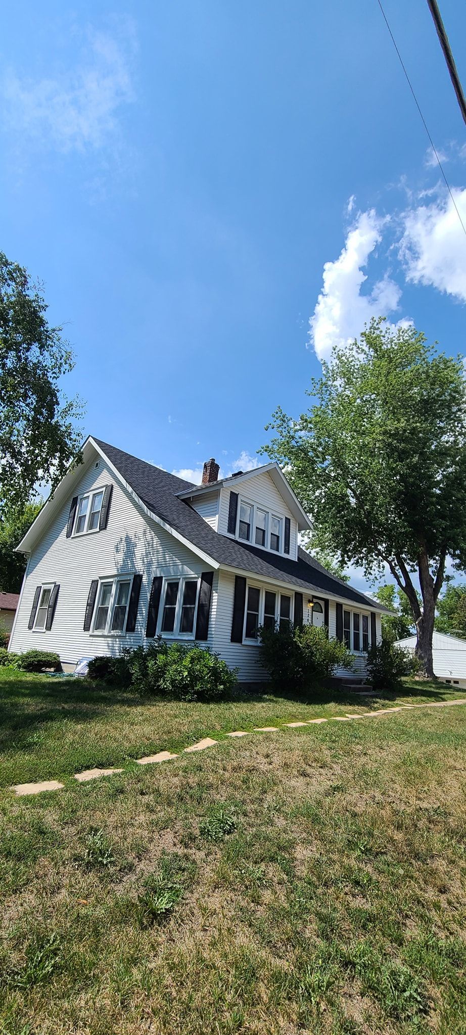 A two-story white house with black shutters stands beneath a bright blue sky on a sunny day.