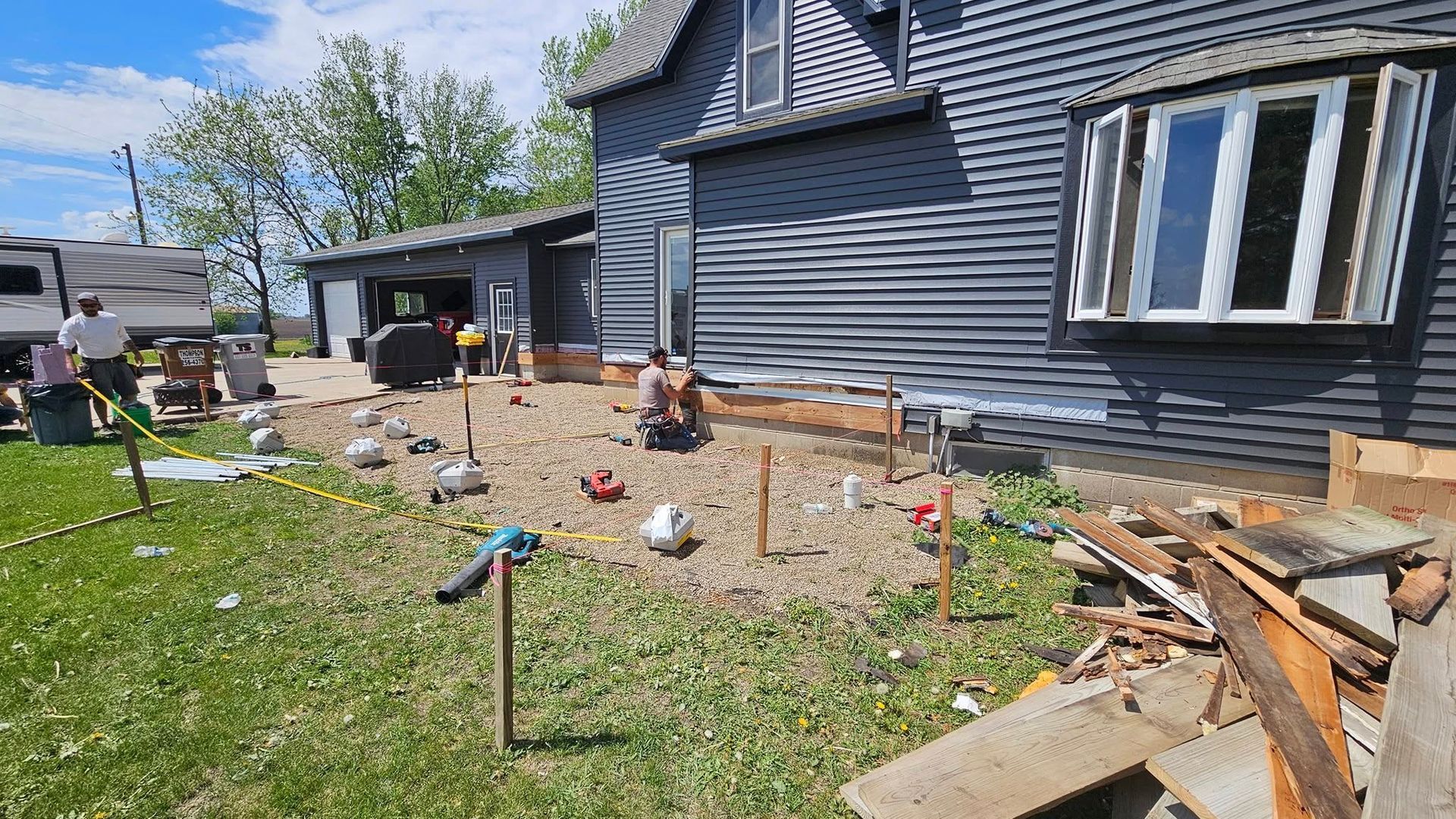 Construction site near a dark gray house. Workers and tools are present, with wooden frames set.