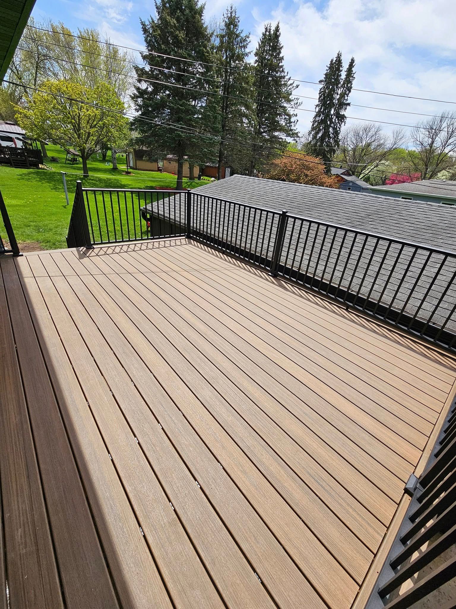 Wooden deck with black railings overlooks green lawn and trees on a sunny day.