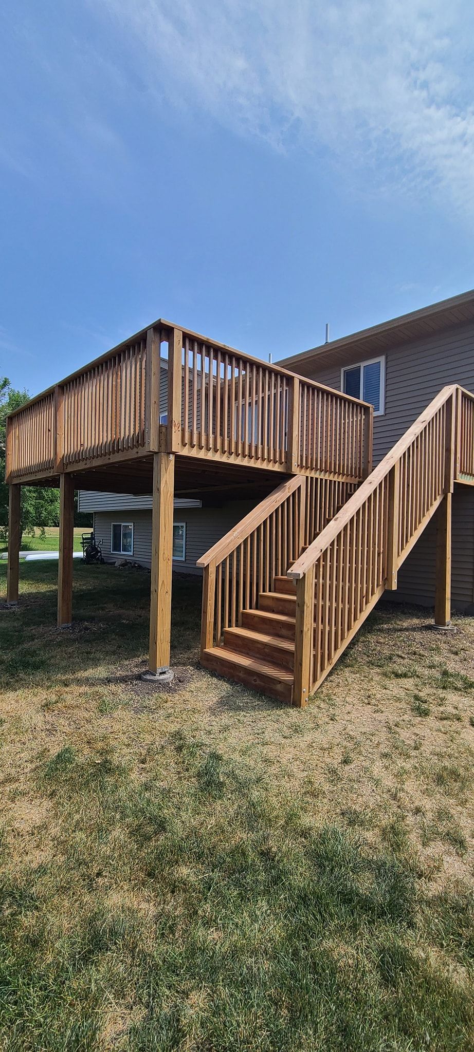 Wooden deck with stairs attached to a two-story house, set on a grassy lawn under a clear blue sky.