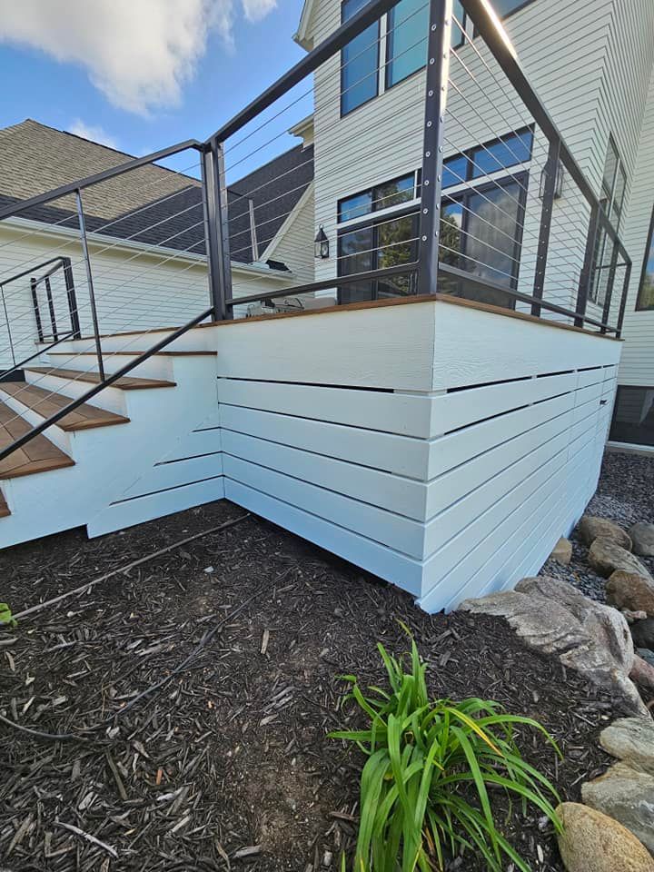 Light blue-sided deck with a black railing, stairs, and cable wire. Located outside a white house on a sunny day.