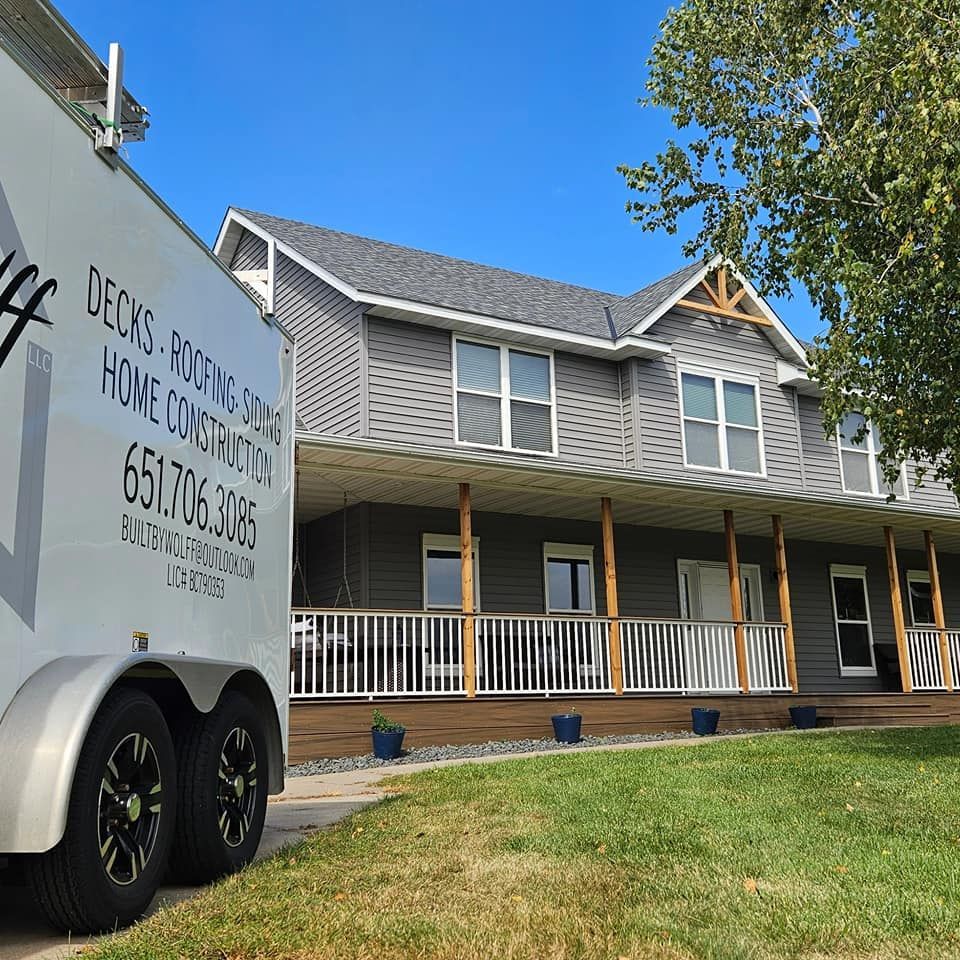 A two-story gray house with a long porch; a trailer with company logo in the foreground.