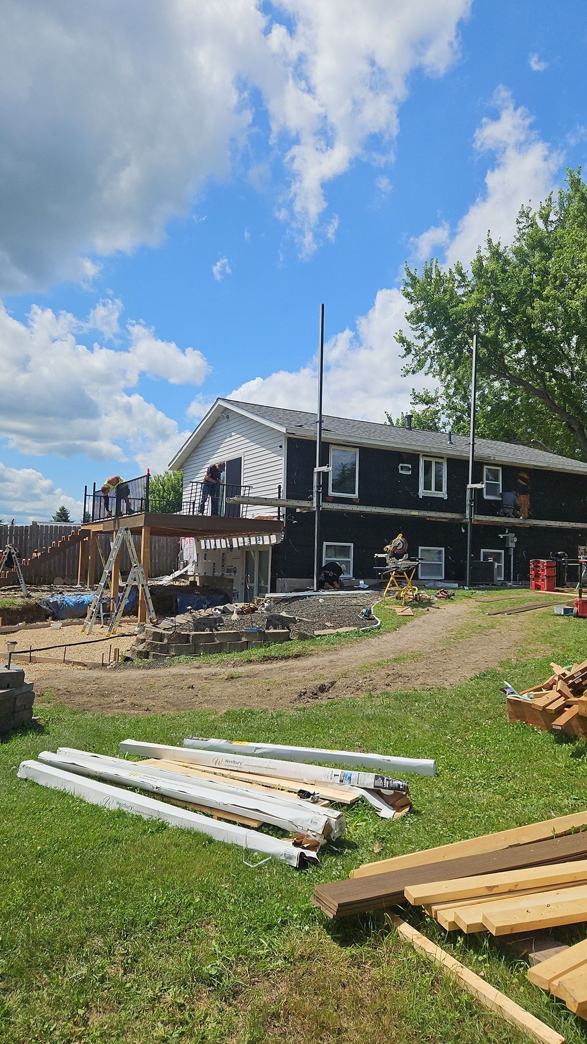 Construction site, a building with black siding, and wood piles. Green grass and blue sky.