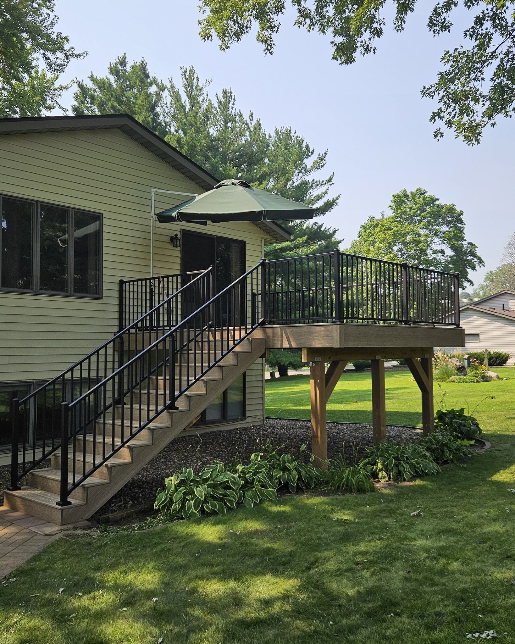 Deck with black railings, stairs, and an umbrella, next to a house with green grass.
