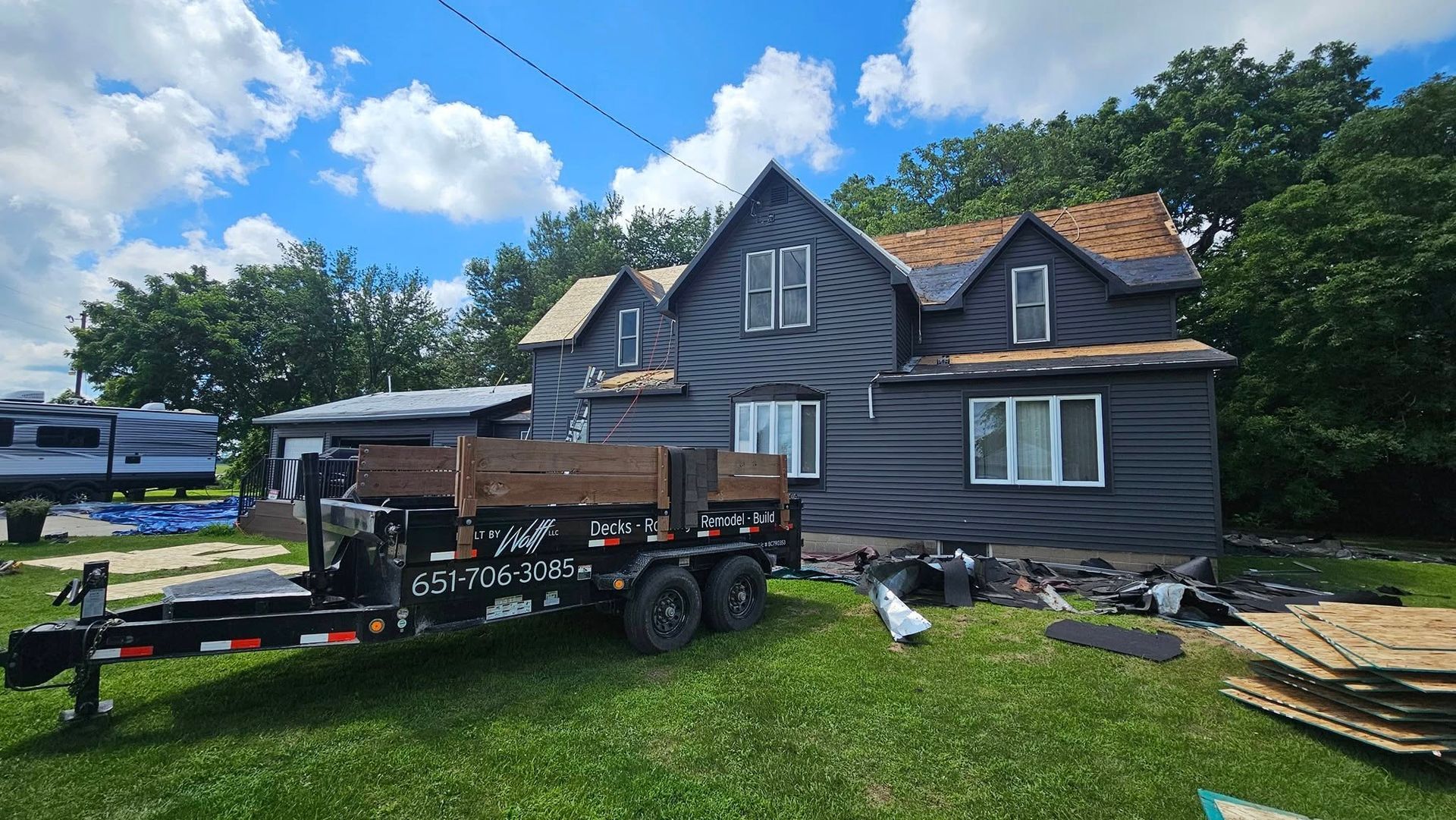 House under construction, black siding, trailer with supplies in yard, sunny day.
