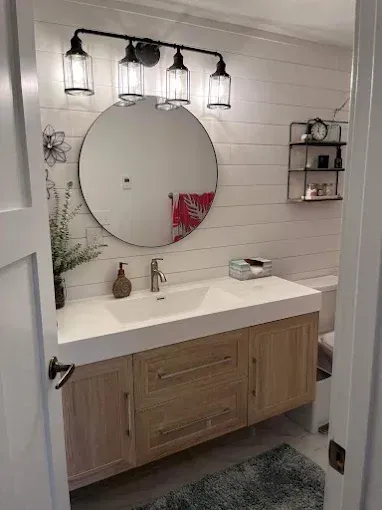 Bathroom with light wood vanity, round mirror, white shiplap walls, and black light fixture.