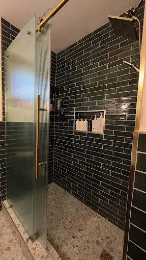 A modern shower with green tiled walls, a glass door, and a pebbled floor.