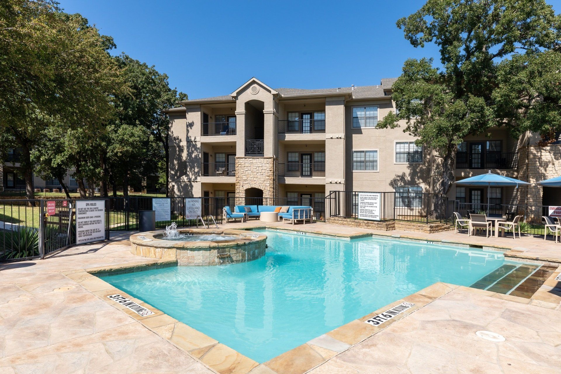 Swimming pool in front of an apartment building, blue water, stone fountain, sunny day, trees.