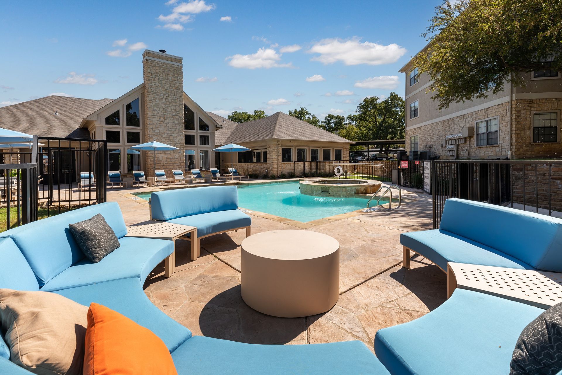 Pool area with blue lounge seating, fountain, and stone buildings under a blue sky.