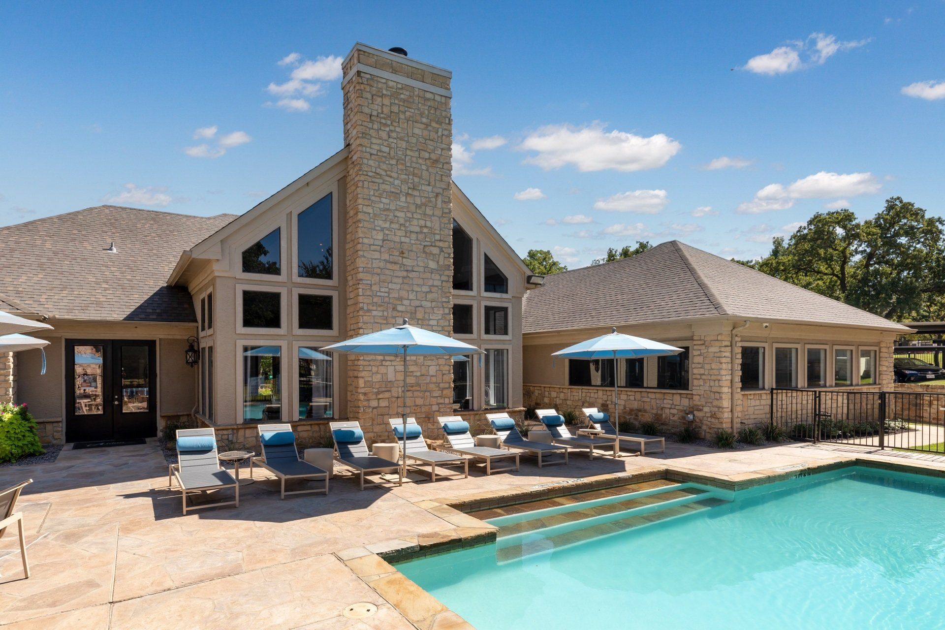 Swimming pool with lounge chairs and umbrellas next to a beige stone building with a tall chimney.