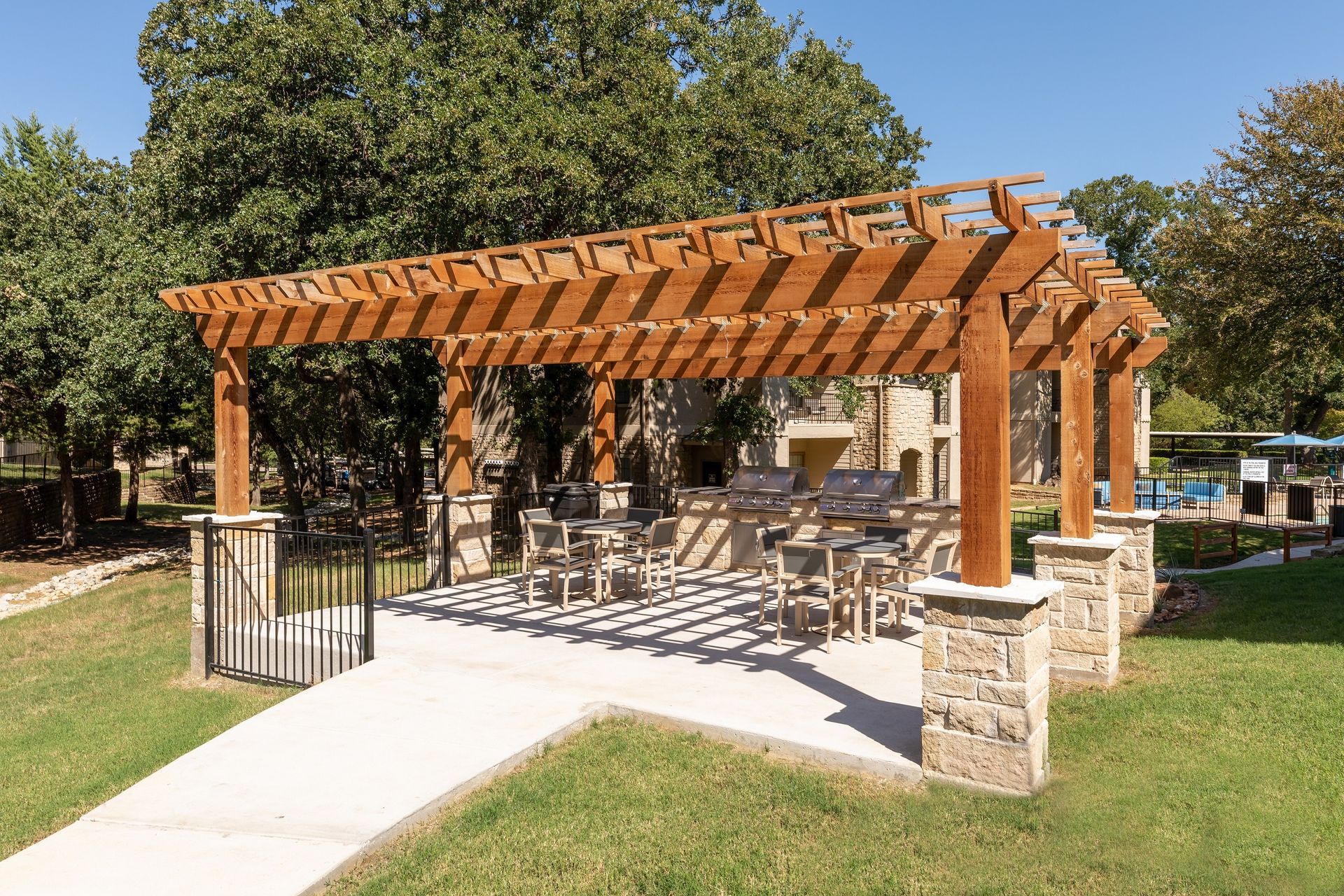 Outdoor wooden pergola with tables, chairs, and built-in grill on a grassy area, with a concrete walkway.
